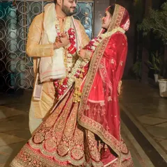 A classic wedding day pose with a candid feel. The couple faces each other, sharing a private moment amidst the celebrations. This shot beautifully captures the bride's flowing red lehenga and the groom's coordinated outfit, highlighting their connection.