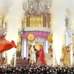 The Jaimala ceremony is elevated with dramatic cold-fire pyrotechnics and smoke effects. The couple stands on a beautifully decorated stage, surrounded by dancers and flags, creating a powerful visual.