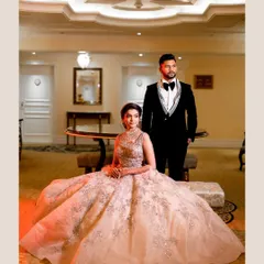 A classic, poised portrait of the couple. The bride is seated, showcasing the full splendor of her gown, while the groom stands beside her, creating a powerful and elegant composition.