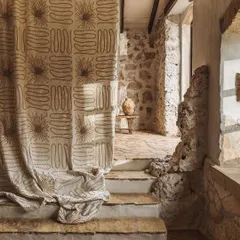 The "Perpétuel Soleil" fabric from Élitis, used as a curtain in a rustic stone interior. The large-scale, hand-beaded embroidery catches the light, transforming the drapery into a piece of textile art that bridges the gap between rustic architecture and refined design.