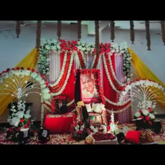 This is a wider view of a satsang stage, showing the complete setup with a red, white, and yellow color scheme. The floor is covered in fresh rose petals, and the sides are decorated with floral stands.