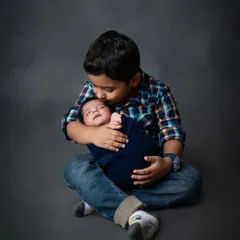 The beginning of a lifelong bond. An older brother lovingly holds and kisses his sleeping newborn sibling, wrapped in a dark blue swaddle.