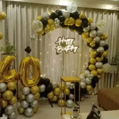 A sophisticated 40th birthday decoration in black, gold, and silver. This setup includes a circular balloon arch, a "Happy Birthday" neon sign, and large foil number balloons.