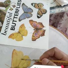 A close-up of a student learning to paint butterflies on fabric, using a reference book for accuracy and detail.