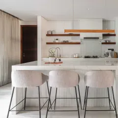 A close-up of the kitchen island, featuring a clean white marble countertop and comfortable upholstered bar stools. Open wooden shelves in the background add warmth and display space.