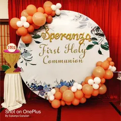 A close-up of the custom backdrop for a First Holy Communion. The design features a floral watercolor print and elegant gold lettering, framed by a delicate arrangement of peach and white balloons.