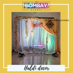 For a softer look, we can create a Haldi backdrop using elegant drapes. This design combines sheer white and rich yellow fabric, accented with a floral border and hanging elements for a classic and bright feel.