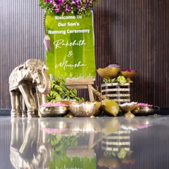 A personalized welcome sign for the naming ceremony, placed on a banana leaf and easel. This is complemented by a brass elephant statue and bowls of fresh flower petals for guests.