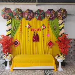 This Haldi decor combines traditional elements with a touch of green. We used a bright yellow backdrop with hanging puppets and colorful tassels, lined with decorative umbrellas and flanked by floral arrangements on white pedestals.