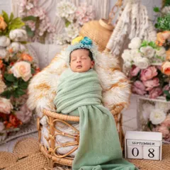 A newborn wrapped in a soft green blanket rests in a boho-style wicker chair, surrounded by pampas grass and muted floral tones. This setup is perfect for families who love a natural, earthy, and chic aesthetic.