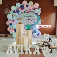 A complete mermaid party setup for Avika's sixth birthday. The decoration includes a pastel balloon arch, themed pedestals, and large light-up letters for the name and 'HBD'.