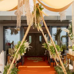 A bohemian entrance that welcomes you with natural elegance. The A-frame bamboo structure is adorned with pampas grass, macrame hangings, and lush greenery, setting a relaxed and chic vibe.