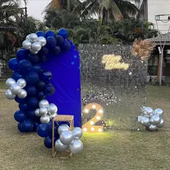A modern outdoor second birthday party setup on a lawn. This decoration combines a royal blue arched backdrop with a silver sequin panel, a neon sign, and a light-up number '2'.