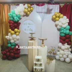 A modern baby shower backdrop with elegant white arched panels and a balloon arrangement in maroon, teal, white, and gold.