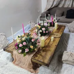 This image shows a rustic wooden low table set for a boho style gathering. The decor includes a textured pink runner, beautiful floral arrangements with pink and yellow roses, and tall, elegant candles on a soft, furry rug.