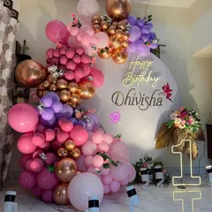 A sophisticated first birthday decoration with a lush, multi-layered balloon arrangement in shades of pink, purple, and rose gold. A light-up number '1' and a custom backdrop add a personal touch.
