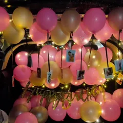 A warm and personal birthday car trunk setup. The space is filled with pink and gold balloons, with small photos hanging from ribbons among them, all illuminated by a "Happy Birthday" banner and soft fairy lights.
