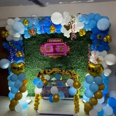 A traditional cradle ceremony decoration for a home. The setup includes a green foliage wall, a decorated cradle, and a blue, white, and gold balloon arch, blending modern and traditional elements.
