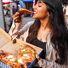 A lifestyle shot of a woman enjoying a slice of pizza in a bustling market setting. This image captures a candid, joyful moment, perfect for a cloud kitchen or takeaway brand wanting to show real-life enjoyment.