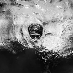 A child's face emerges from the dark water, their eyes wide and captivating in a dramatic and artistic black and white portrait that blurs the line between life and art.