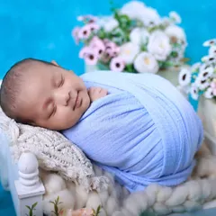 A picture of pure bliss. We are experts at soothing babies and capturing those heart-melting sleepy smiles. This newborn is comfortably swaddled against a soft blue backdrop, surrounded by delicate flowers.