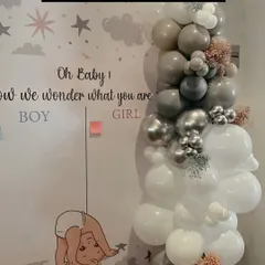 A "Guess The Gender" voting board is a fun activity for a baby shower. This one features a cute design and is placed next to a stylish grey and white balloon garland.