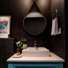 A close-up of the vanity area in the moody bathroom. The combination of the dark wall tiles, the round leather-strapped mirror, and the vibrant art creates a rich, layered look that feels both dramatic and inviting.