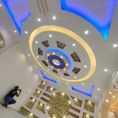 This is an example of a highly decorative, multi-level false ceiling. It combines circular motifs, intricate CNC-cut patterns, and dual-color LED lighting to create a grand and luxurious effect.