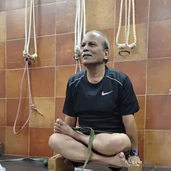Seated in Padmasana (Lotus Pose) on blocks with a strap for support. This setup helps in training the hips and knees for the full pose while maintaining an upright and comfortable spine.