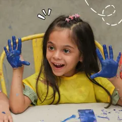 A young girl with paint on her hands looks up with pure excitement. My kids' parties are all about letting them have fun and be creative.
