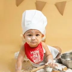 This little baker, with a dusting of flour on his nose and a cute "Mom + Dad = Me" bib, crawls towards the camera, ready for his next kitchen adventure.