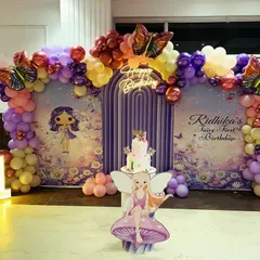 A whimsical 'Fairy First Birthday' setup featuring a custom backdrop with fairy illustrations, butterfly accents, and a pastel balloon garland in purple, pink, and yellow.