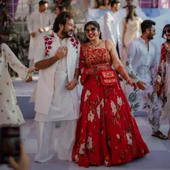 The bride and groom make their entrance at the welcome party. Her vibrant red floral lehenga and his coordinated white kurta with a floral embroidered jacket create a stunning couple's look that is both festive and fashionable.