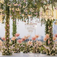 A frontal view of the open-air mandap, showcasing the harmony between the lush floral pillars, the whimsical oversized paper flowers, and the urban cityscape in the distance. This design blends nature with modern romance.