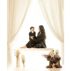 A wider view of the same intimate studio moment. The composition, with its beautiful floral arrangement and soft drapery, creates an elegant and timeless portrait of a mother and her child sharing a special connection.