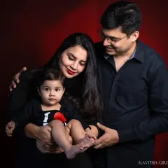 A warm and intimate family studio portrait. The deep red background adds a touch of richness, while the family's loving interaction remains the central focus.