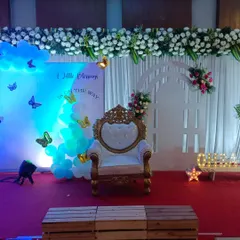 This baby shower stage combines modern and traditional touches. It has an elegant throne chair, a white floral garland overhead, and a backdrop with blue lighting and butterfly accents to welcome the new baby.