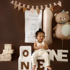A joyful first birthday photo with a warm, brown-toned teddy bear theme. The setup includes a large "1" balloon, gift boxes, and a happy baby boy.