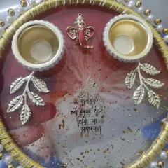 Here is a closer look at a custom pooja thali, perfect for your home mandir. This design features a rich red and white resin base with gold flakes, a Gayatri Mantra, a small Ganesha, and holders for roli and chawal, all bordered with delicate pearls.