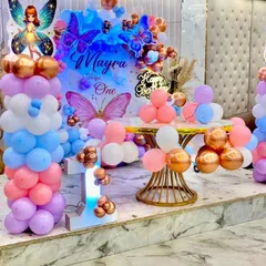 A magical butterfly-themed first birthday stage decoration. The setup includes a personalized backdrop for "Mayra," pastel balloon pillars with fairy cutouts, and a beautifully decorated cake table.