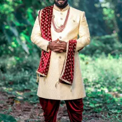 A happy client looking absolutely majestic for his outdoor wedding ceremony. He is wearing a custom-tailored cream sherwani with a maroon velvet stole, paired with traditional dhoti pants and red shoes.