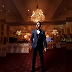 A dapper groom standing tall in a grand ballroom. The low-angle shot emphasizes his sharp tuxedo and the magnificent chandeliers above.