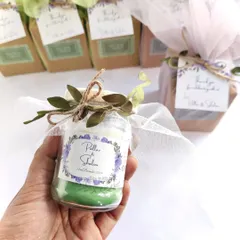 A hand holding a fully packaged wedding favor, a green layered candle with a custom "Pillai & Shalini" sticker, white net wrap, and leafy tie.