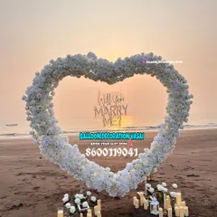 A close-up of the "Will You Marry Me?" proposal setup at sunset. The heart-shaped arch is made of dense white flowers, with the neon sign glowing warmly as the sun goes down, creating a magical and picturesque scene.