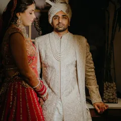 A classic wedding portrait of the couple in their regal attire. The groom looks towards the camera while the bride stands by his side, creating a formal yet intimate image that highlights their stunning outfits.