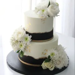 A timeless black, white, and gold engagement cake. This two-tiered design features a torn-edge effect and is adorned with fresh white florals for a modern and sophisticated look.