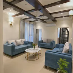 A wider view of the Doon Homestay living room, showing how the space comes together. The arrangement of the teal blue sofas, nested marble-top tables, and soft curtains creates a comfortable and stylish area for guests to relax.