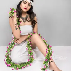 A seated portrait from the 'Gaia' series. The model is dressed in white with a floral garland, looking directly at the camera with a powerful yet gentle expression, capturing the essence of a mythical goddess.