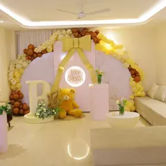 A wide view of an elegant baby shower setup in a living room. The decor features a large circular backdrop with a golden bow, a beautiful balloon arch in neutral tones, and a light-up sign for "Baby Singh."