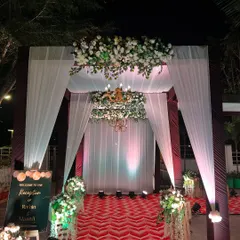 A classic and grand reception entrance. The walkway is marked by a red chevron carpet, leading to an elegant structure draped in white fabric and adorned with white floral arrangements and a chandelier.